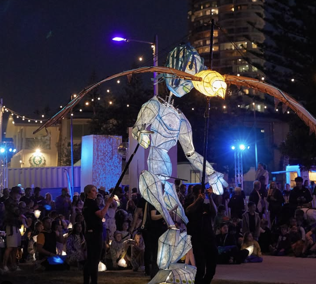 An outdoor night scene with a lit-up, large, illuminated puppet of a person with wings, being held up by several people, surrounded by a crowd of spectators.
