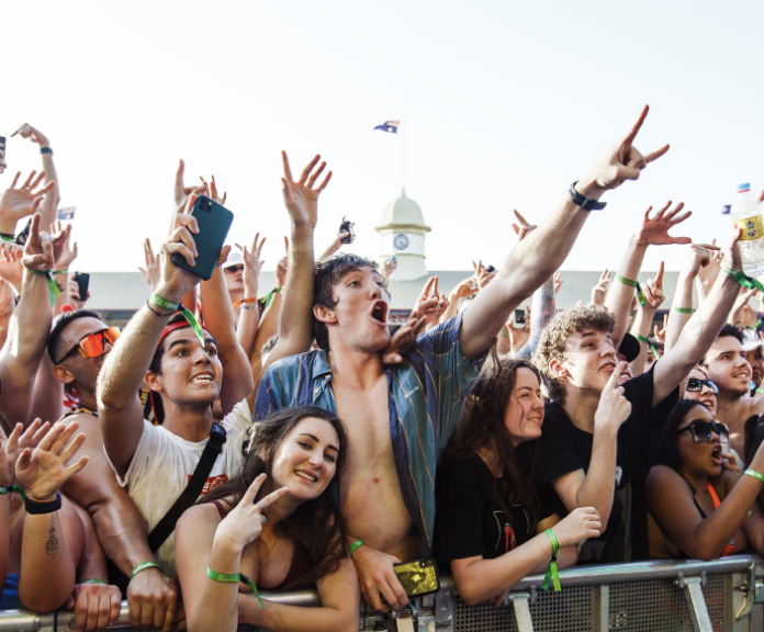 Group of young people at an outdoor concert or festival, cheering and taking photos, with some raising their hands and smiling.