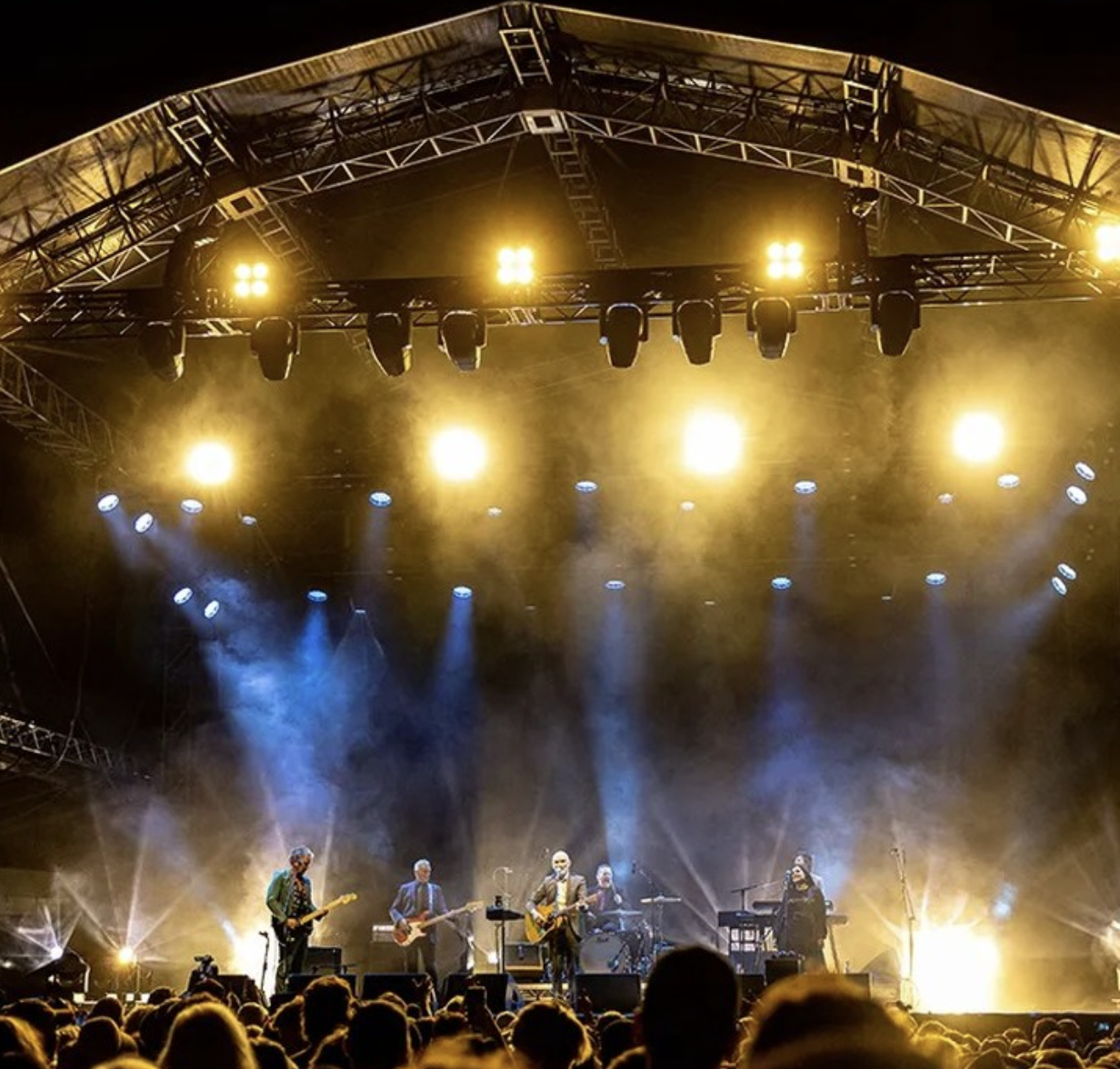 Live concert on an outdoor stage with a band performing, bright yellow and white stage lights, and an audience in the foreground.