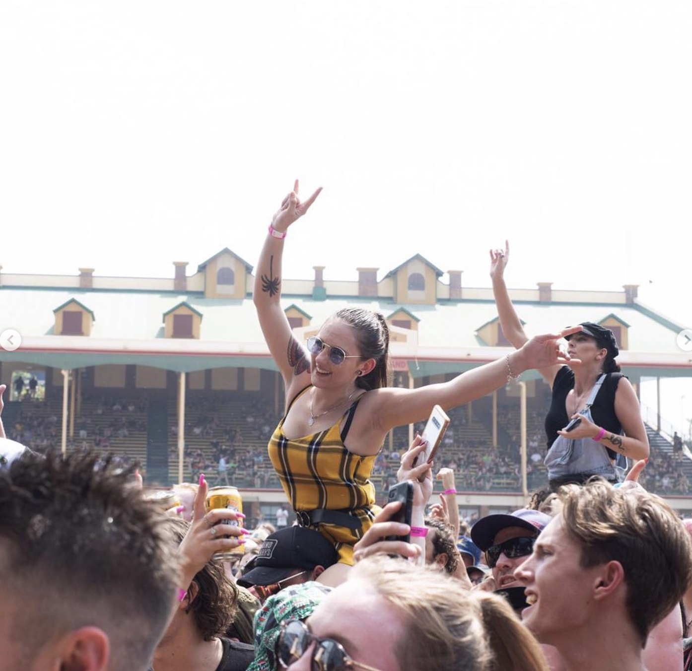 People at an outdoor concert crowd surfing, with a woman in the center raising her arms and smiling, holding a drink. The background features a building with a distinctive roof and multiple levels of seating.