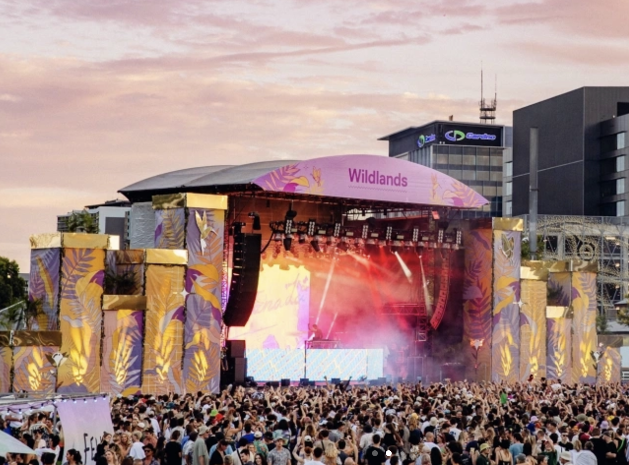 A large outdoor concert stage at sunset with a crowd of people in front. The stage is decorated with tropical leaf patterns and has a pink banner that reads 'Wildlands'. Smoke effects and bright lighting are visible on the stage. Buildings are seen in the background.