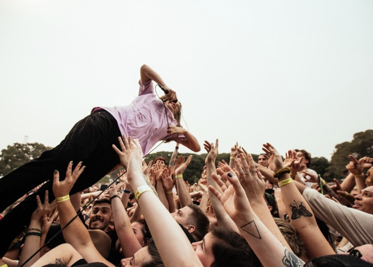 A singer performs at an outdoor concert, crowd reaching up to touch or hold him, with the singer lying on the hands of audience members.
