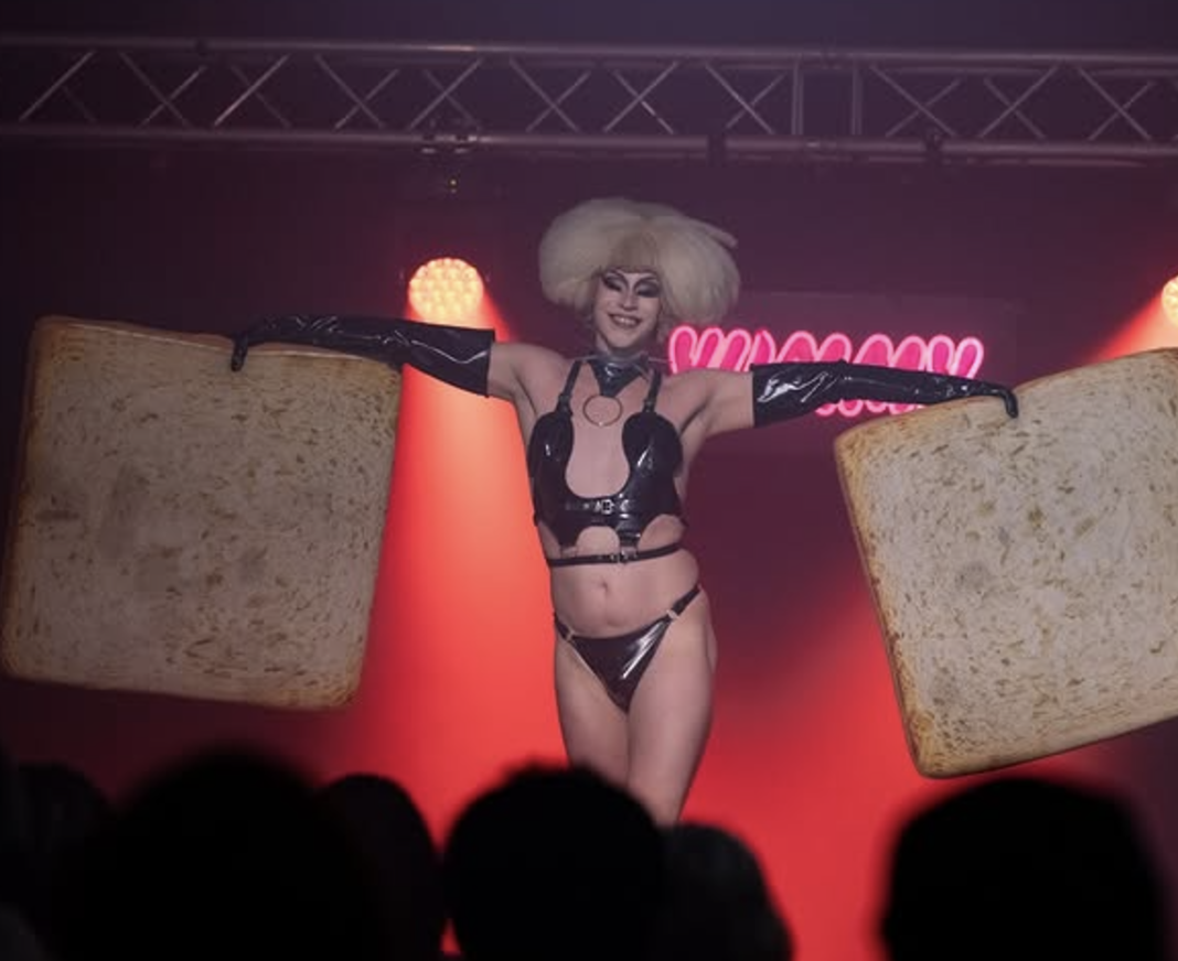 Performer on stage with large toasted bread props, wearing black latex outfit and gloves, blonde wig, in front of a red spotlight background and pink neon sign.