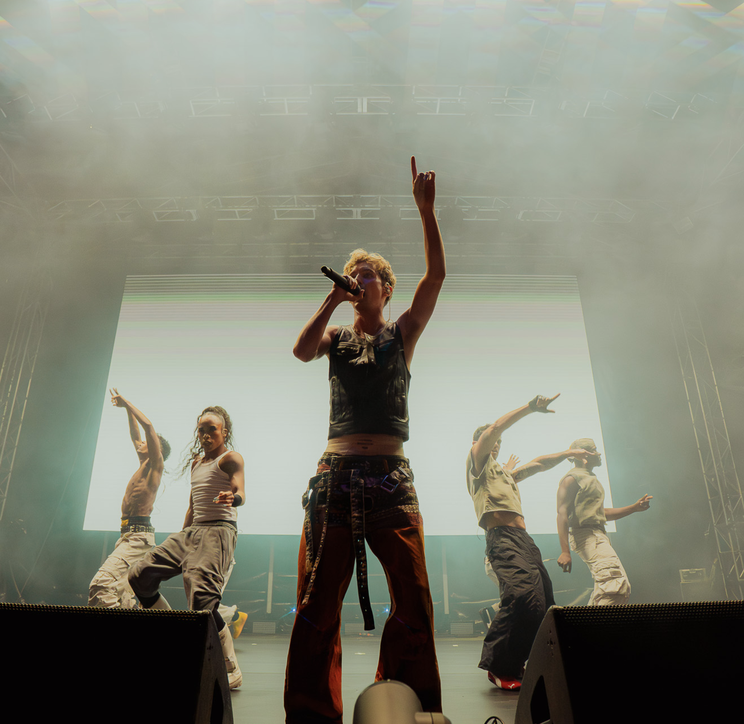 A female singer performing on stage with four backup dancers, standing in front, holding a microphone, with a large screen behind them displaying light green, blue, and pink horizontal lines, and stage lighting creating a smoky atmosphere.