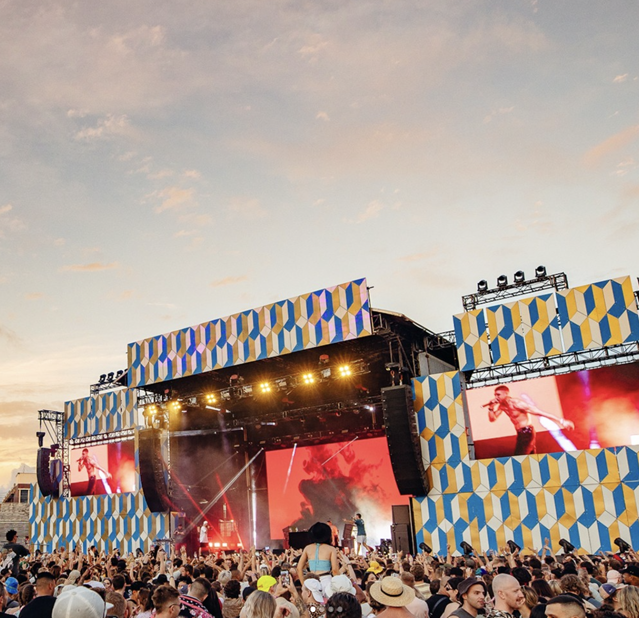 Outdoor music festival with a large crowd, stage, and big screen, during sunset, with performers on stage and decorative geometric patterned banners.