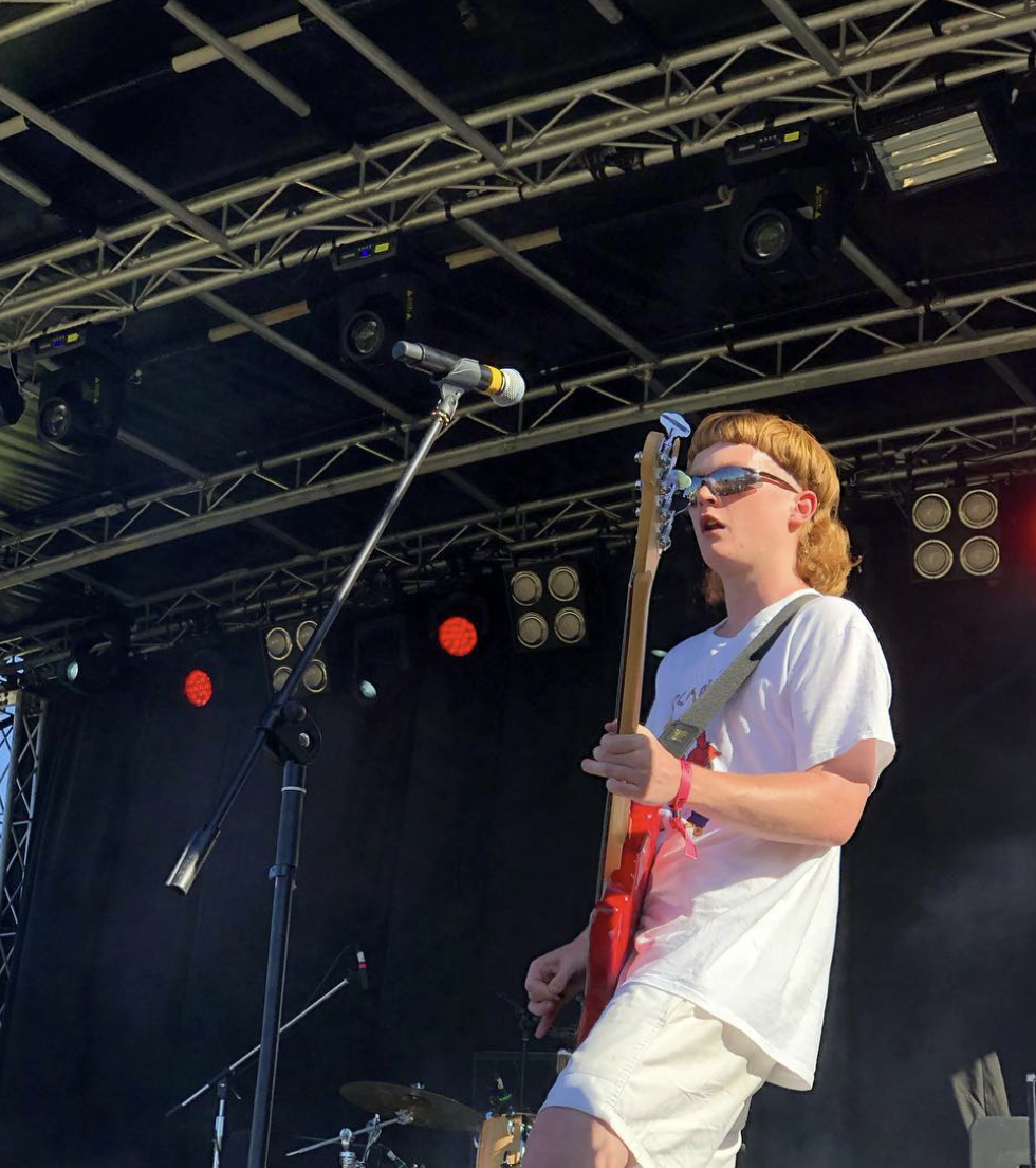 A young man with red hair, sunglasses, and a white T-shirt playing an electric guitar on stage during a live performance with stage lights and sound equipment overhead.