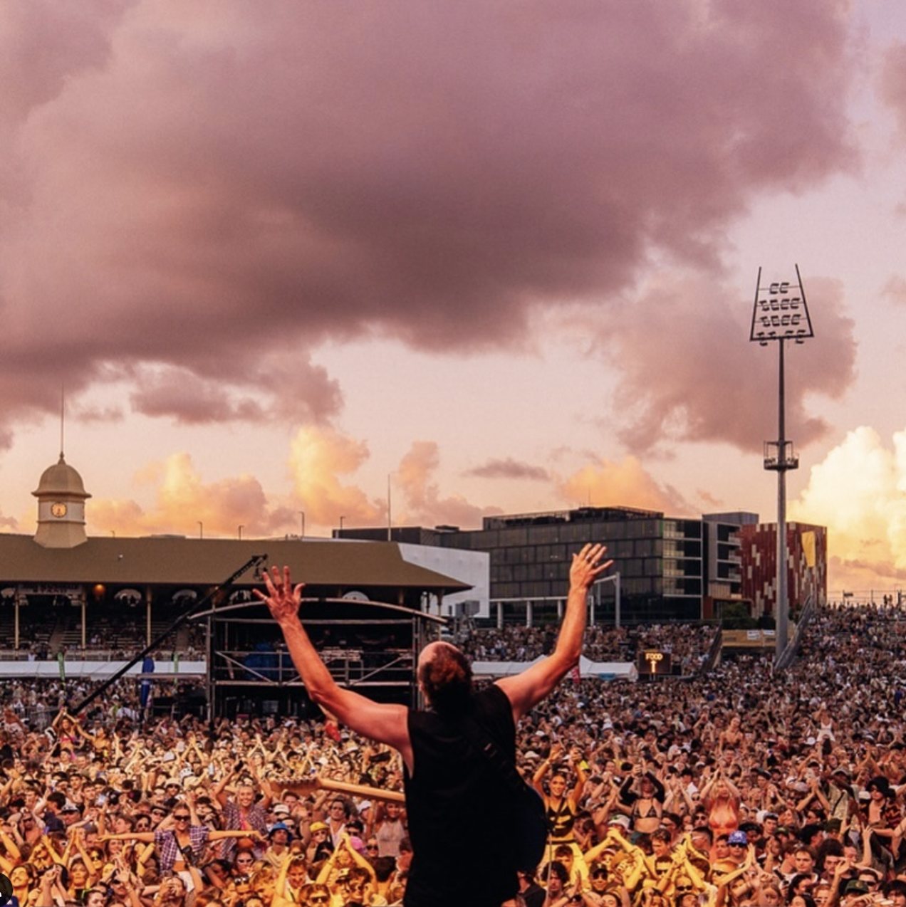 Concert crowd with performer on stage raising arms, stadium lights, modern buildings, and a sunset sky with clouds.
