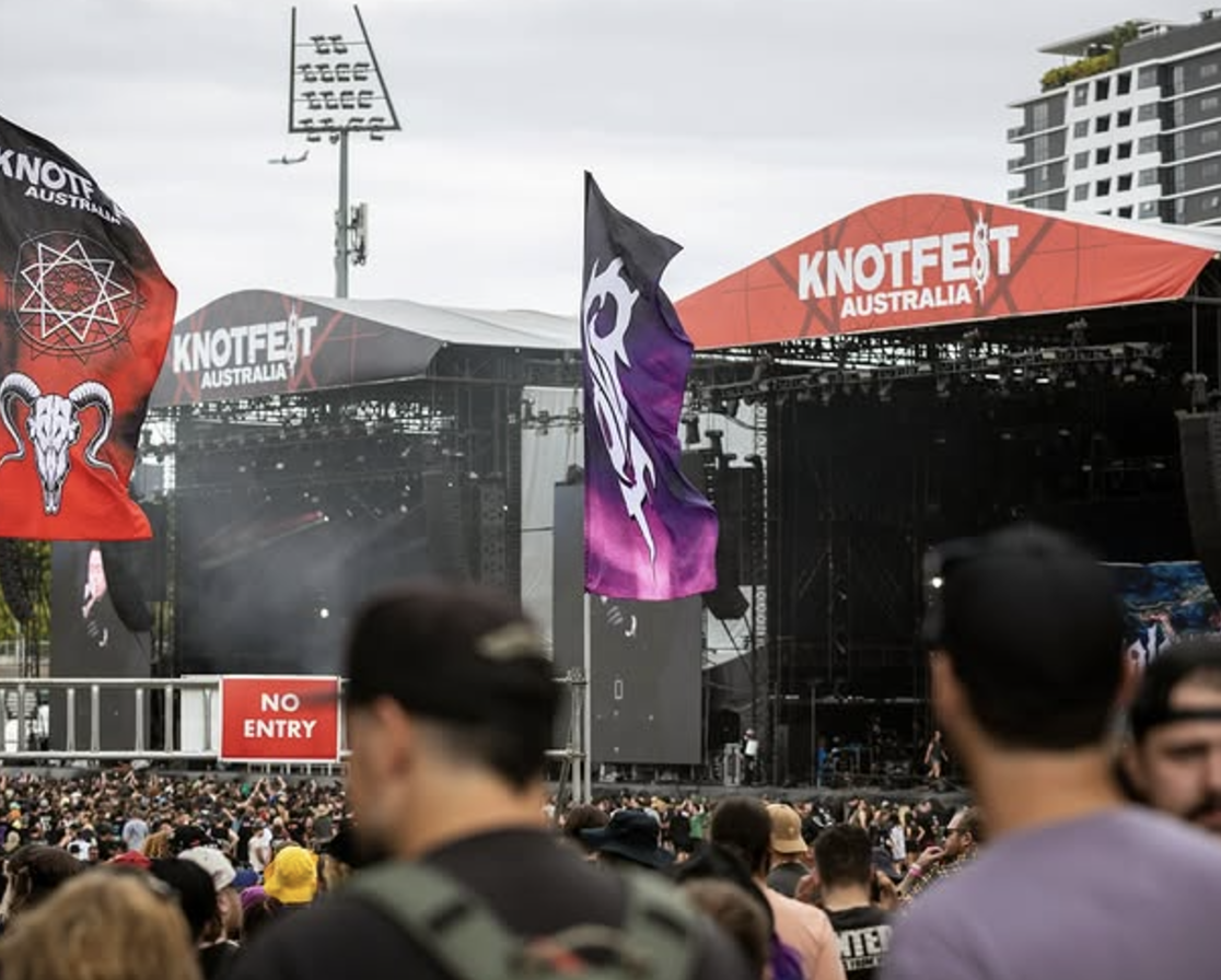 A large crowd at an outdoor music festival with a stage labeled 'Knockfest Australia.' Flags with artistic designs are hanging above the crowd, and there is a 'No Entry' sign near the stage.
