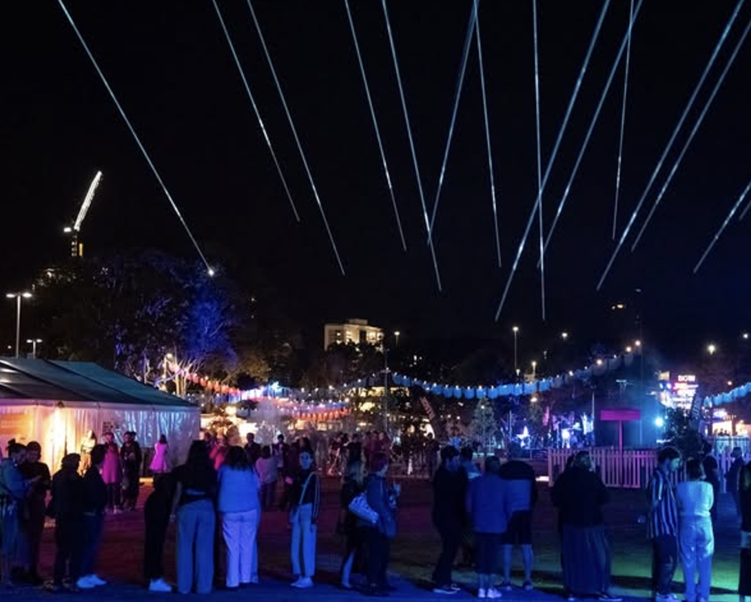 Nighttime outdoor festival with colorful lights, a large crowd of people, tents, and fireworks or sky lanterns in the sky.
