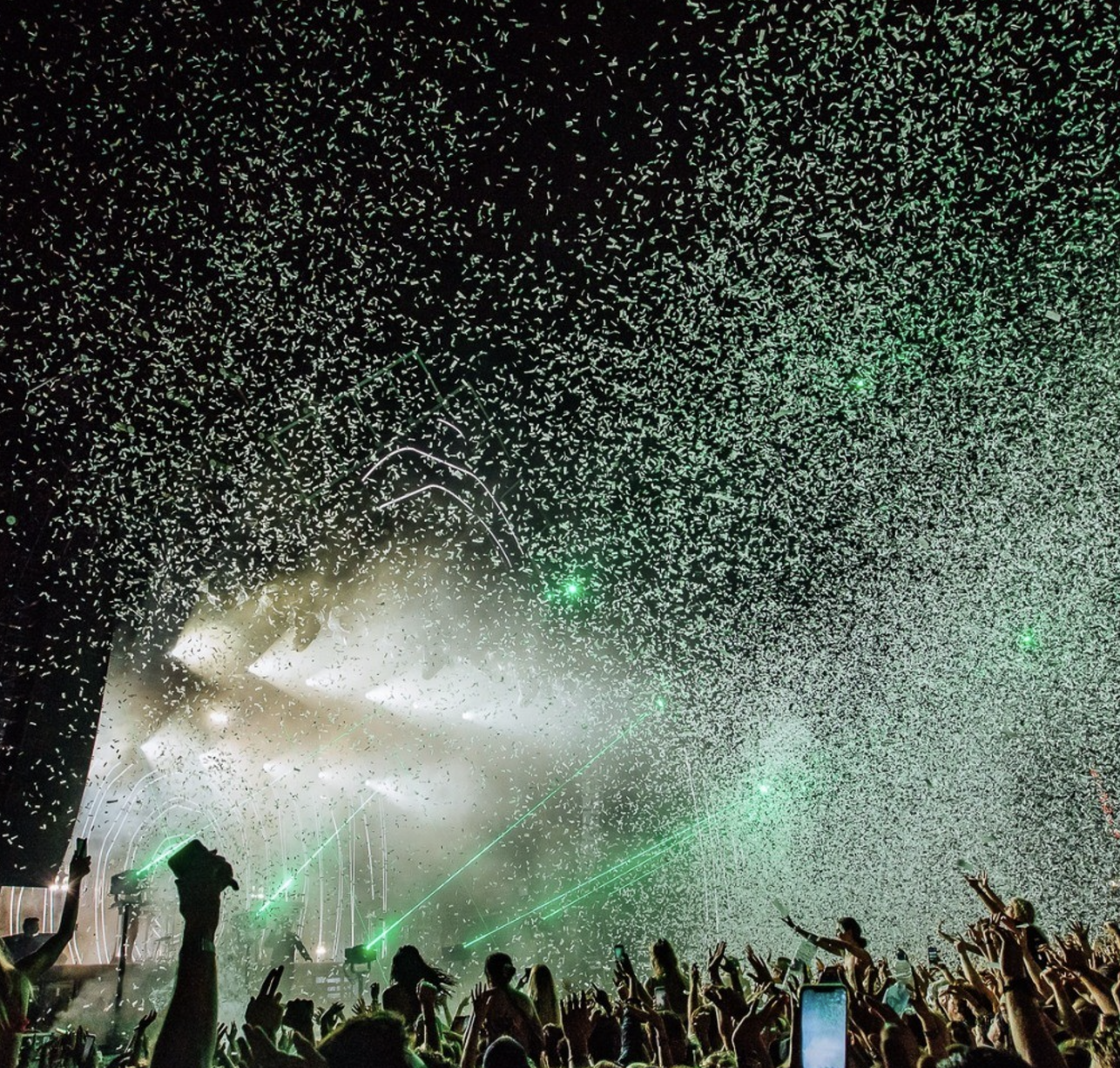 Crowd at a concert or festival with confetti and laser lights in the air, and people raising their hands, some taking photos.