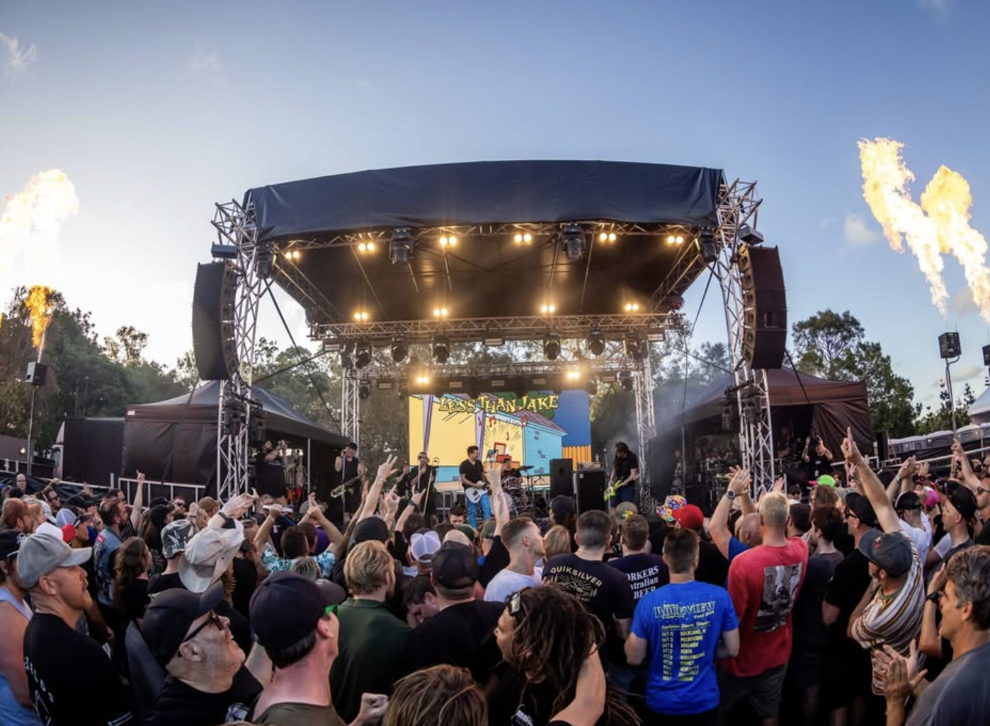 A live outdoor concert with a band performing on stage and an enthusiastic crowd cheering, during sunset or early evening.