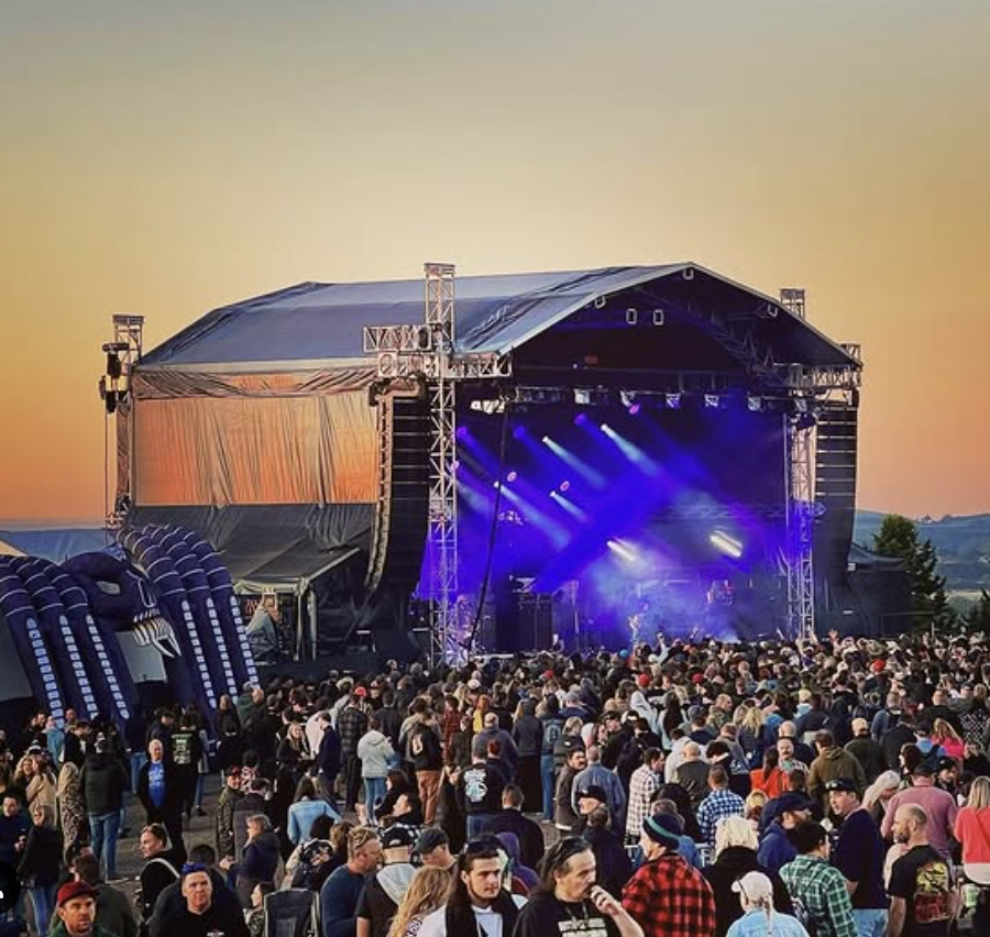 Crowd gathering at an outdoor concert at sunset with a large stage and colorful lighting.