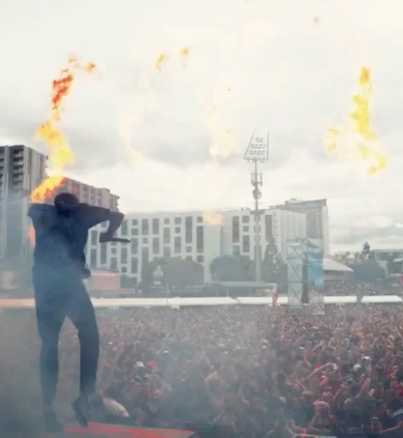 Person on stage holding a flaming torch at a crowded outdoor event with buildings and a cloudy sky in the background.