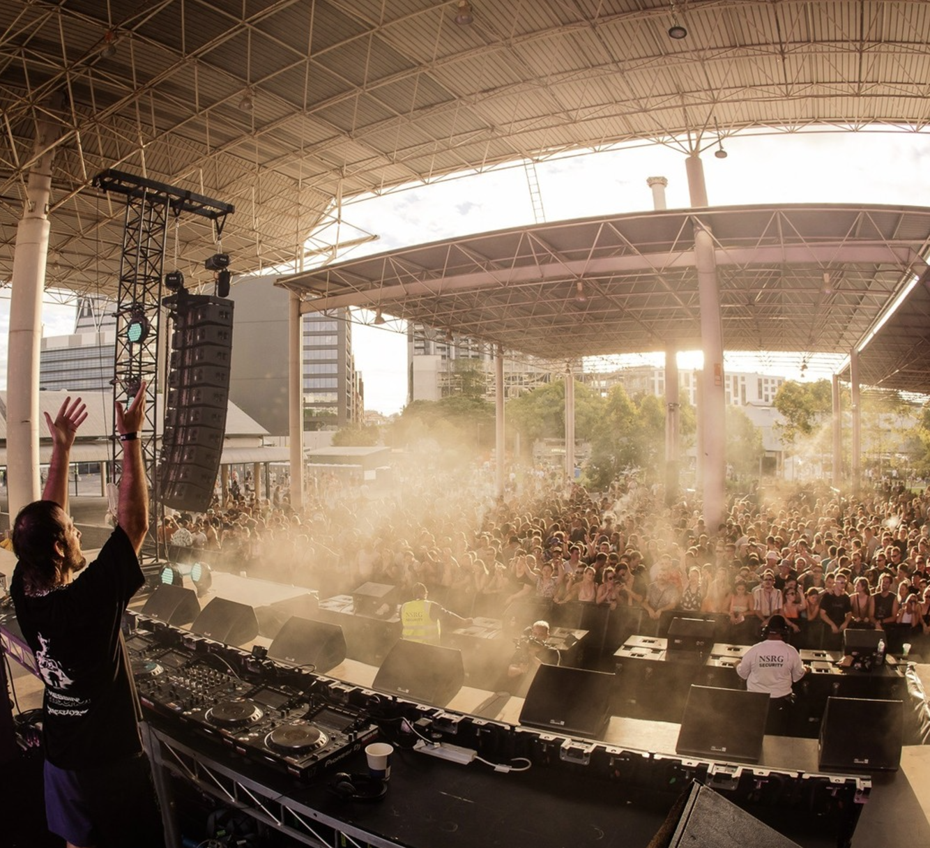 A DJ performs on stage at an outdoor concert, raising his hands while facing a large crowd under a modern, open-air pavilion with city buildings in the background, during sunset.