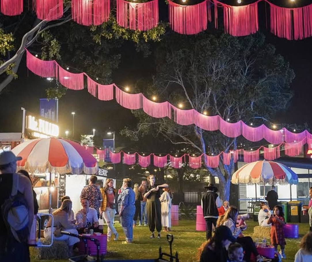 Night scene at a fair or festival with pink hanging decorations, colorful umbrellas, and people enjoying various activities.