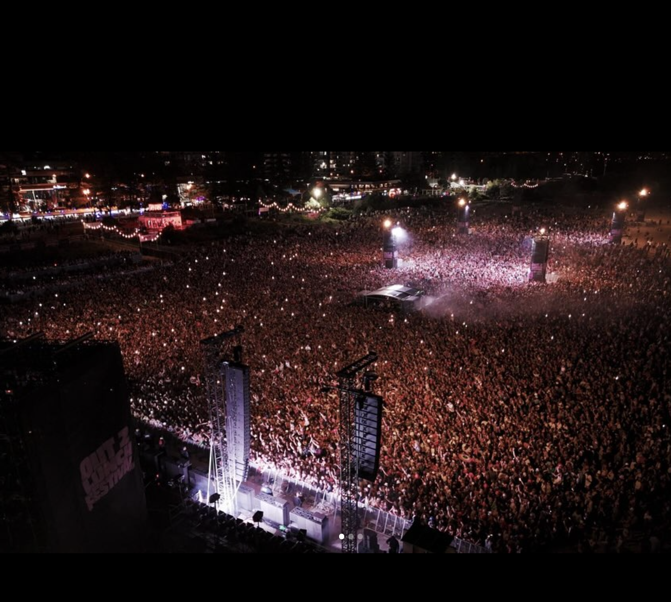 A crowded outdoor music concert at night with bright stage lights, large speakers, and a full audience.