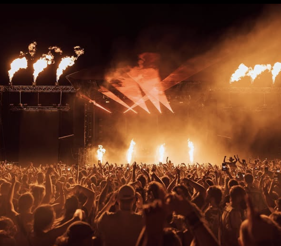A large crowd at a concert or music festival with fiery pyrotechnics and bright lighting on stage at night.