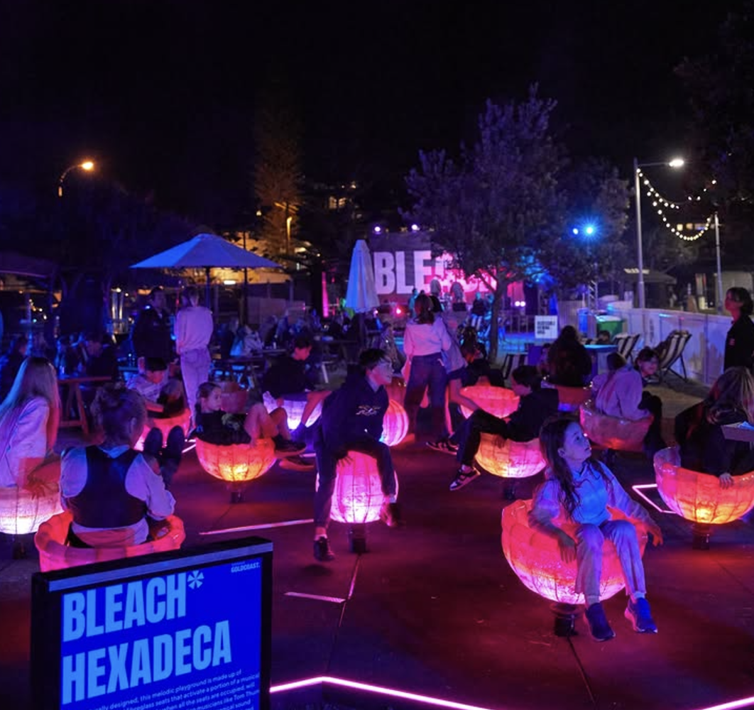 People at a nighttime outdoor event with colorful illuminated bowls, a stage with a sign that says 'BLEACH HEXADECA,' and string lights overhead.