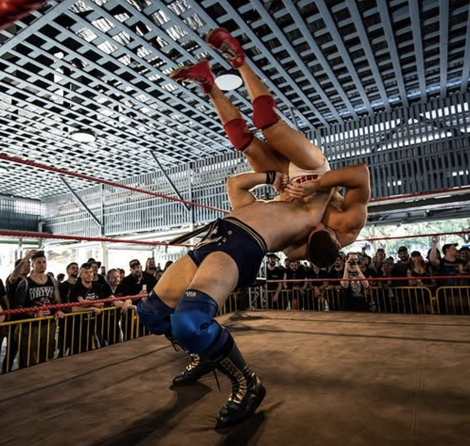 Two male wrestlers engaged in a wrestling match inside a ring with ropes. One wrestler is upside down, and the other is flipping him. An audience watches from outside the ring in a large, open space with a metal-patterned ceiling.