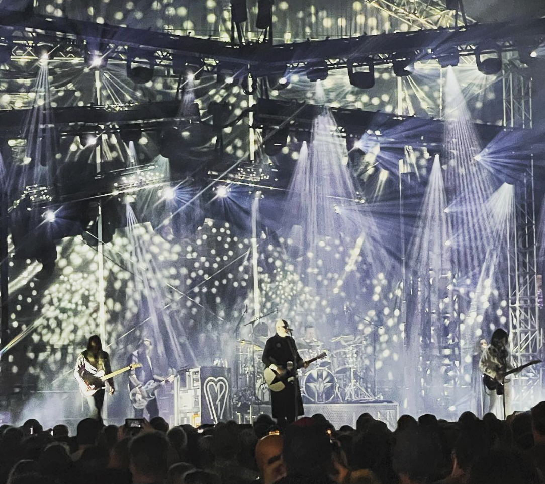 A live band performs on stage with dramatic lighting, including spotlights and patterned effects, while an audience watches from below.