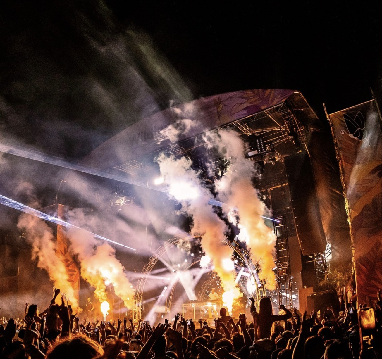 A nighttime outdoor concert with a stage, bright lights, smoke effects, and a crowd of people enjoying the performance.