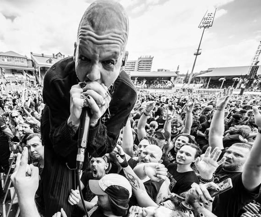 A black and white photo of a man with a shaved head singing into a microphone at a crowded outdoor concert or gathering, with many people raising their hands and cheering in the background.