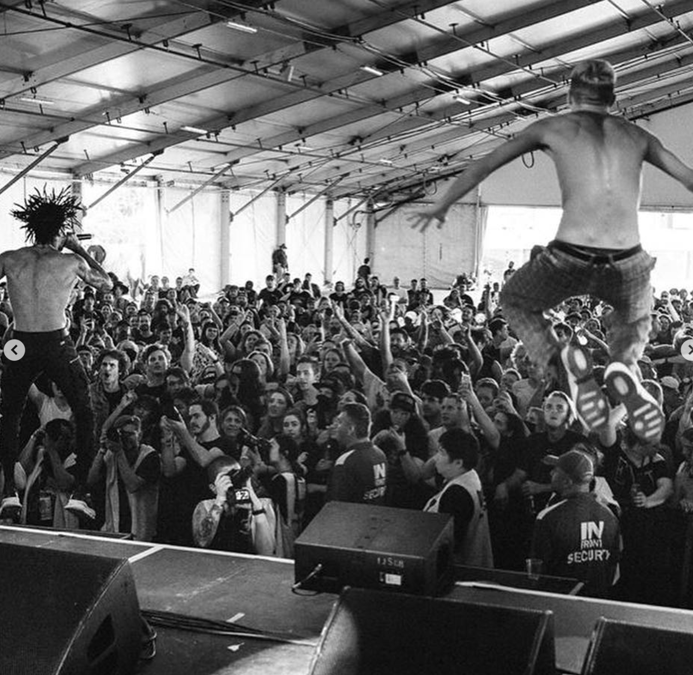 A black and white photograph of a concert or event inside a large tent or gymnasium. The stage is at the front, with two shirtless performers, one with a microphone and spiky hair, and the other mid-jump or dance with arms outstretched. The crowd is tightly packed, with many raising their hands and cheering. Security is visible at the front near the stage.
