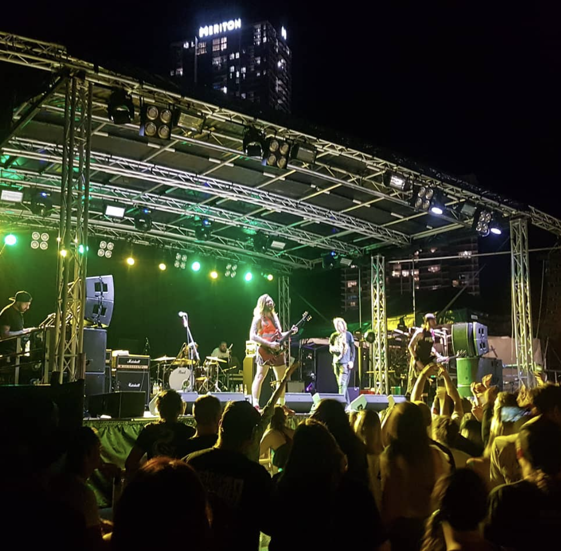A live music concert at night with a band performing on an outdoor stage illuminated by green and yellow lights, audience in the foreground, and tall buildings in the background.