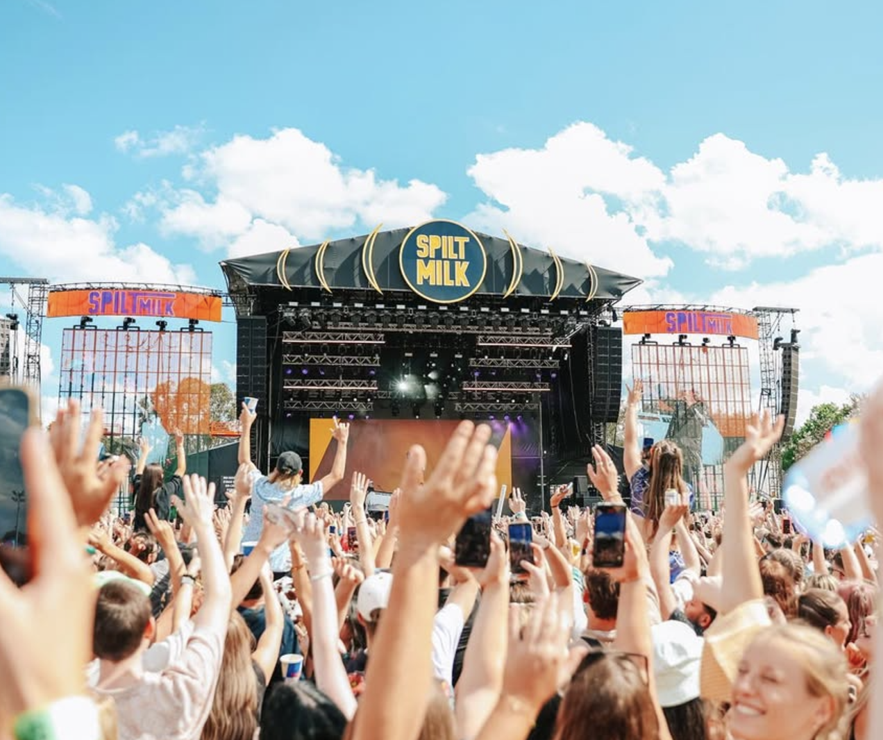 Crowd at outdoor music festival with a stage labeled "Split Milk" under a partly cloudy sky.