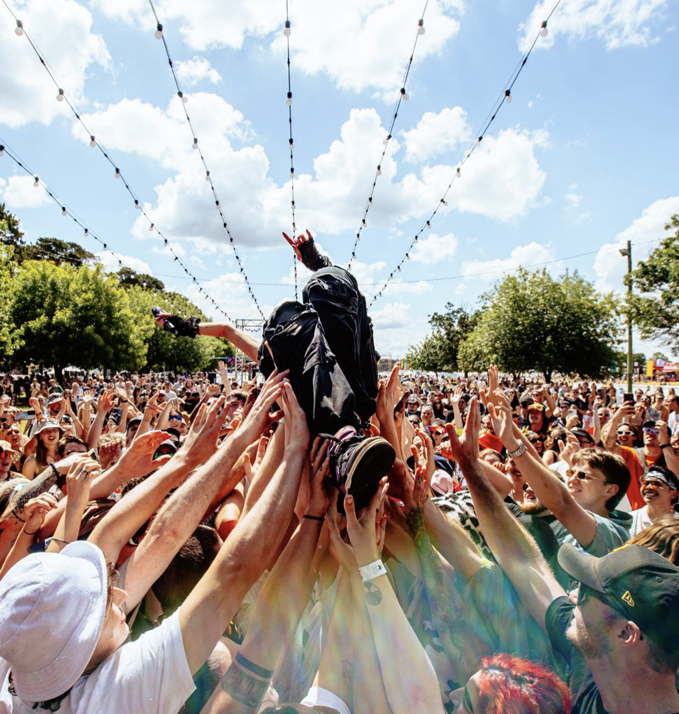 Crowd of people outdoors at a festival or concert, with a person being lifted above the crowd, reaching towards the sky, under strings of lights and a partly cloudy sky.