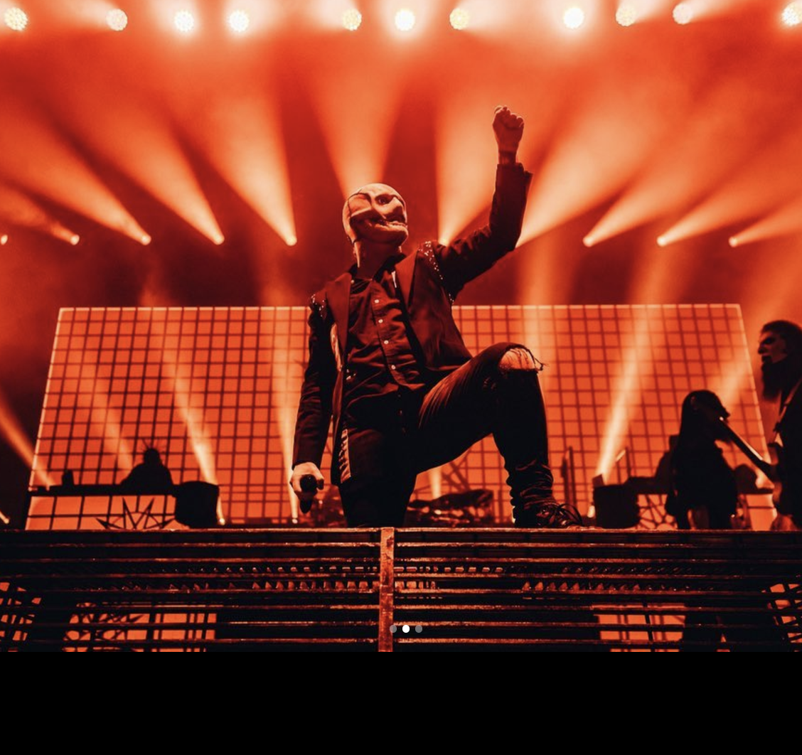 Performer on stage with a microphone, wearing a mask and raising a fist, illuminated by orange stage lights with a backdrop of large illuminated panels.