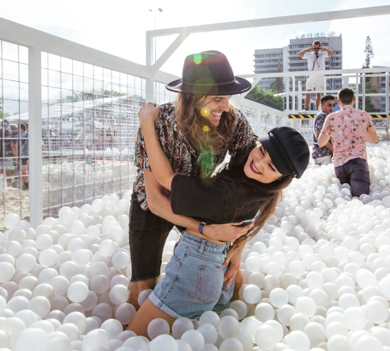 Two women playfully hugging and laughing in a ball pit filled with white balls at an outdoor event, with other people and structures in the background.