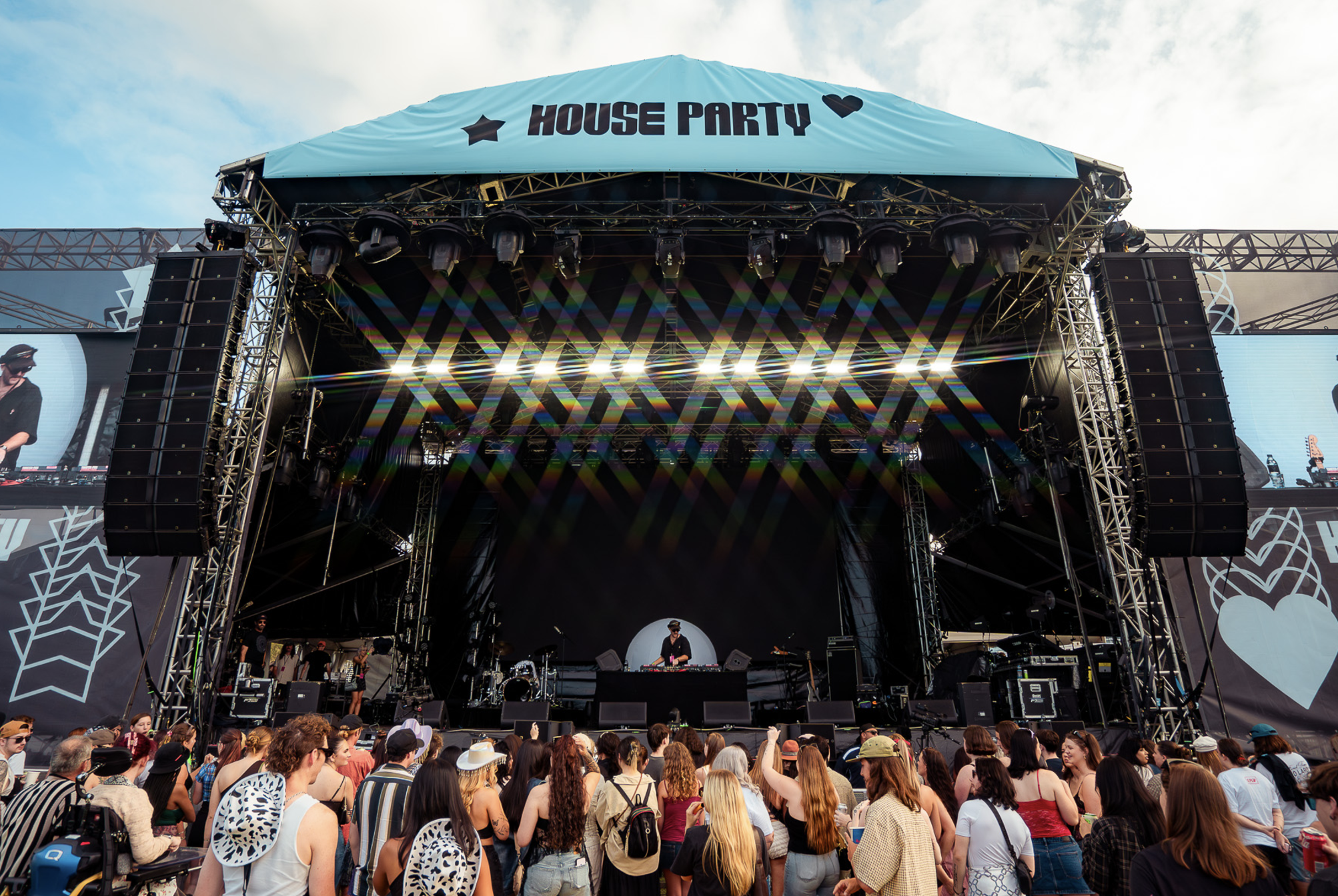 Crowd at an outdoor house party music concert with large stage, DJ, and colorful LED lights, under a blue sky.