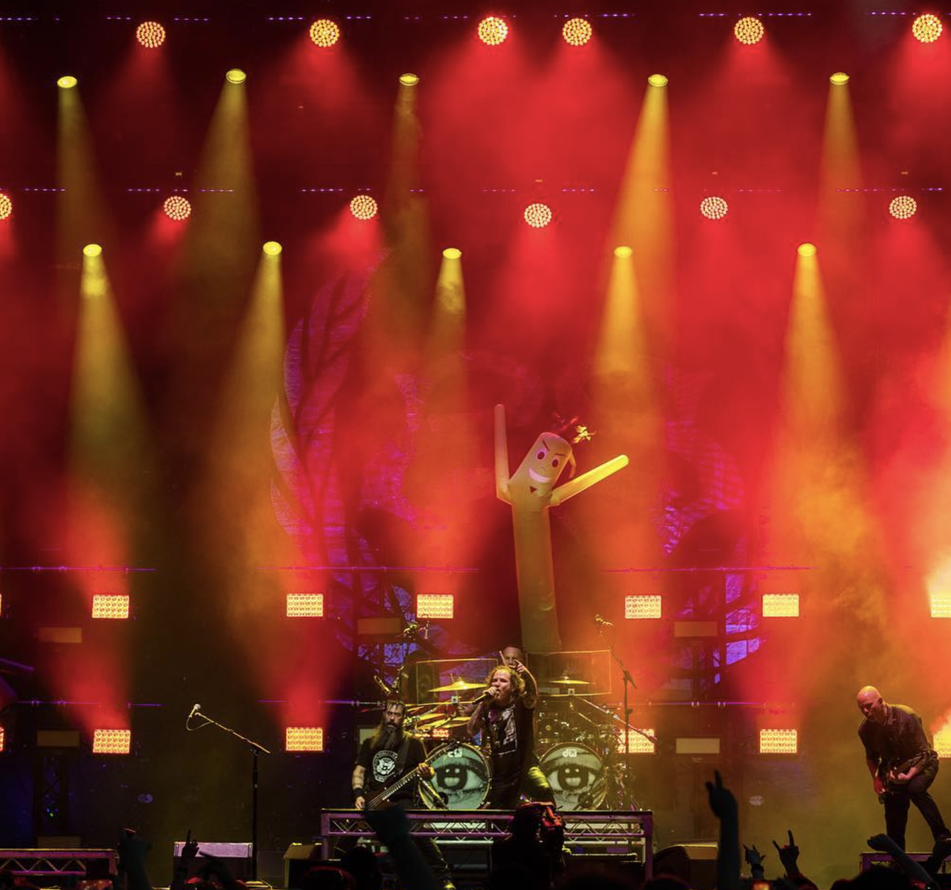 A live concert stage with yellow, orange, and red lighting, featuring a band performing with a large inflatable cactus with face and arms in the background.