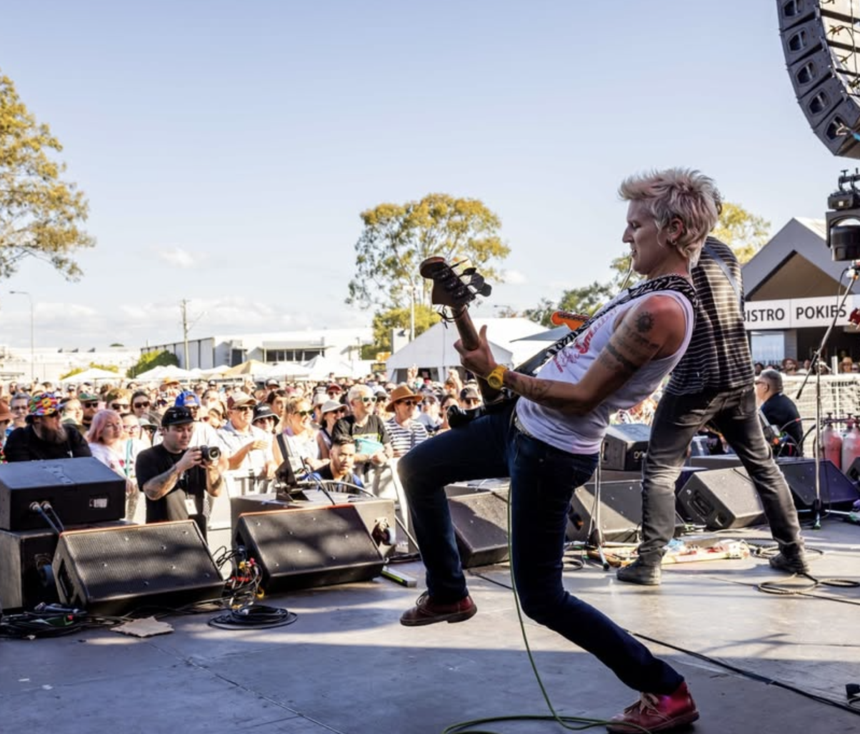 Musician with pink hair performing guitar on stage at an outdoor concert with crowd of fans watching.