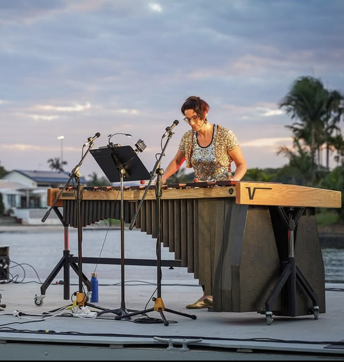 A woman playing a marimba outdoors during sunset with trees and houses in the background.