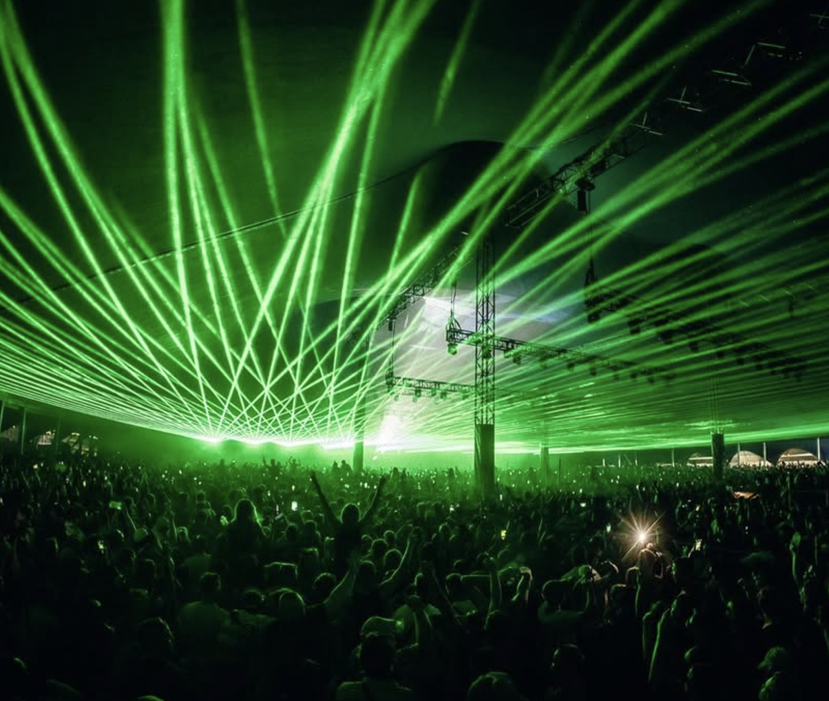 A large crowd at a concert or music festival with green laser lights illuminating the venue and audience, and a stage with lighting rig.