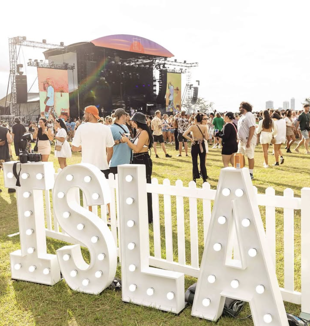 Outdoor music festival at sunset with a stage, large crowd, and illuminated letters spelling 'ISLA' in the foreground.
