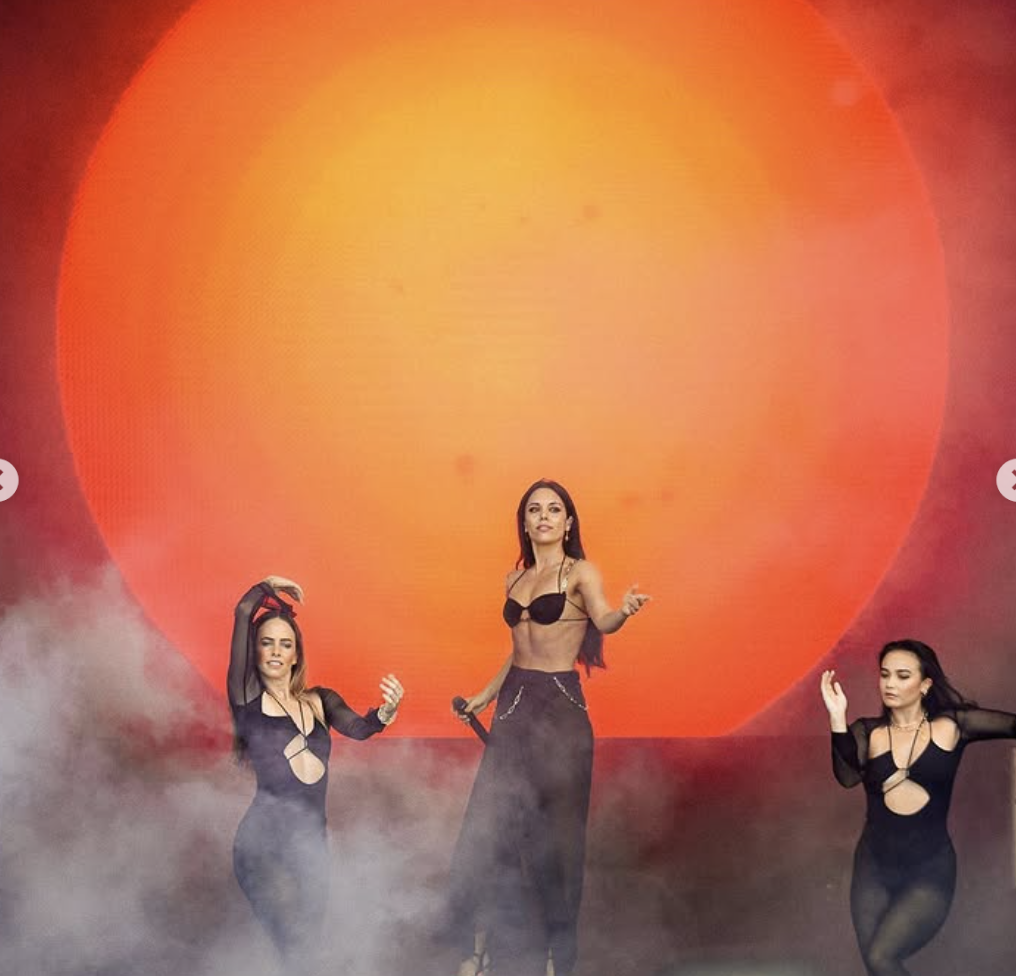 Three women in black outfits performing on stage with a large orange and yellow sun backdrop and fog effects.