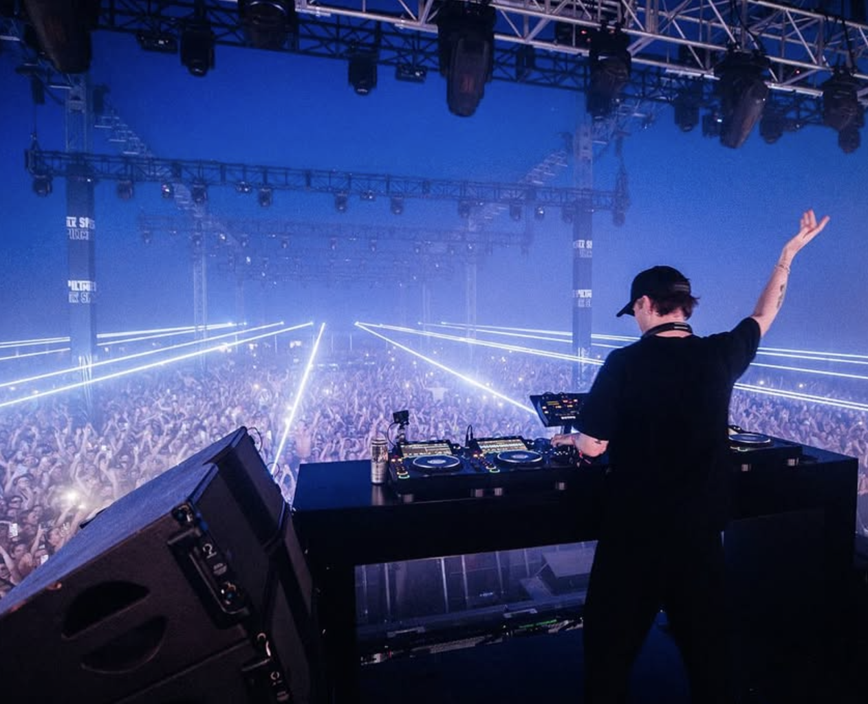 A DJ performs on stage at a concert with a large crowd of festival attendees, laser lights, and stage equipment.