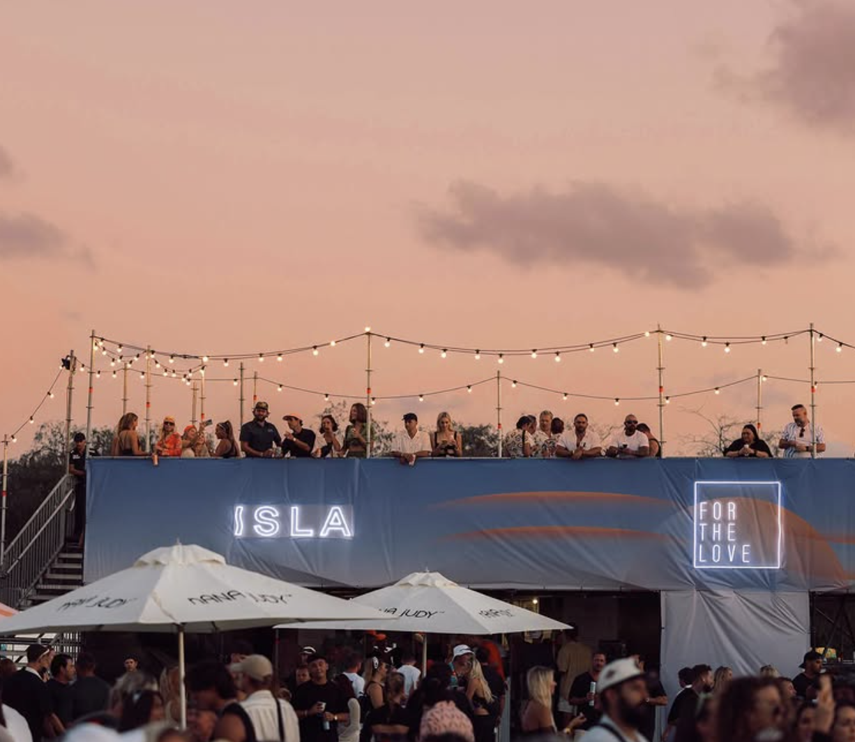 People gathered at an outdoor event during sunset with string lights and neon signs that say 'ISLA' and 'FOR THE LOVE.' There are umbrellas and a large crowd of attendees.