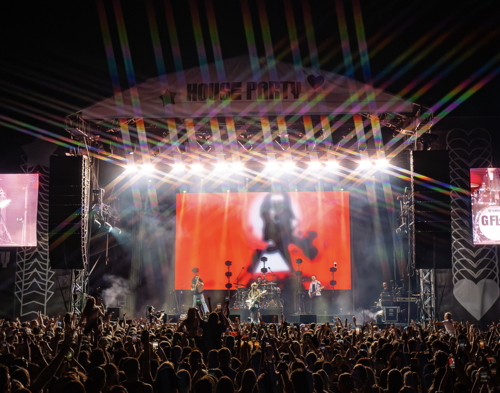 A large concert stage at night with bright lights, performers, and a crowd of people with their hands raised.