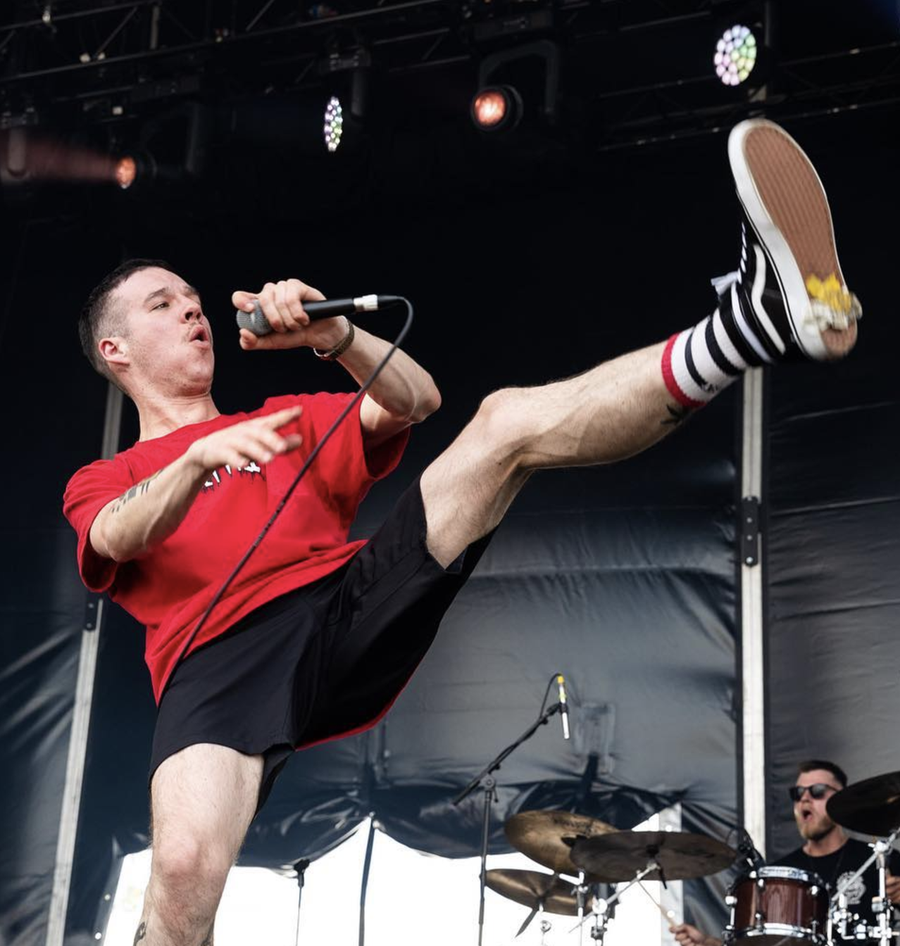 A male singer wearing a red shirt, black shorts, and striped socks performing with a microphone on stage at a concert, raising one leg high with a fierce expression.
