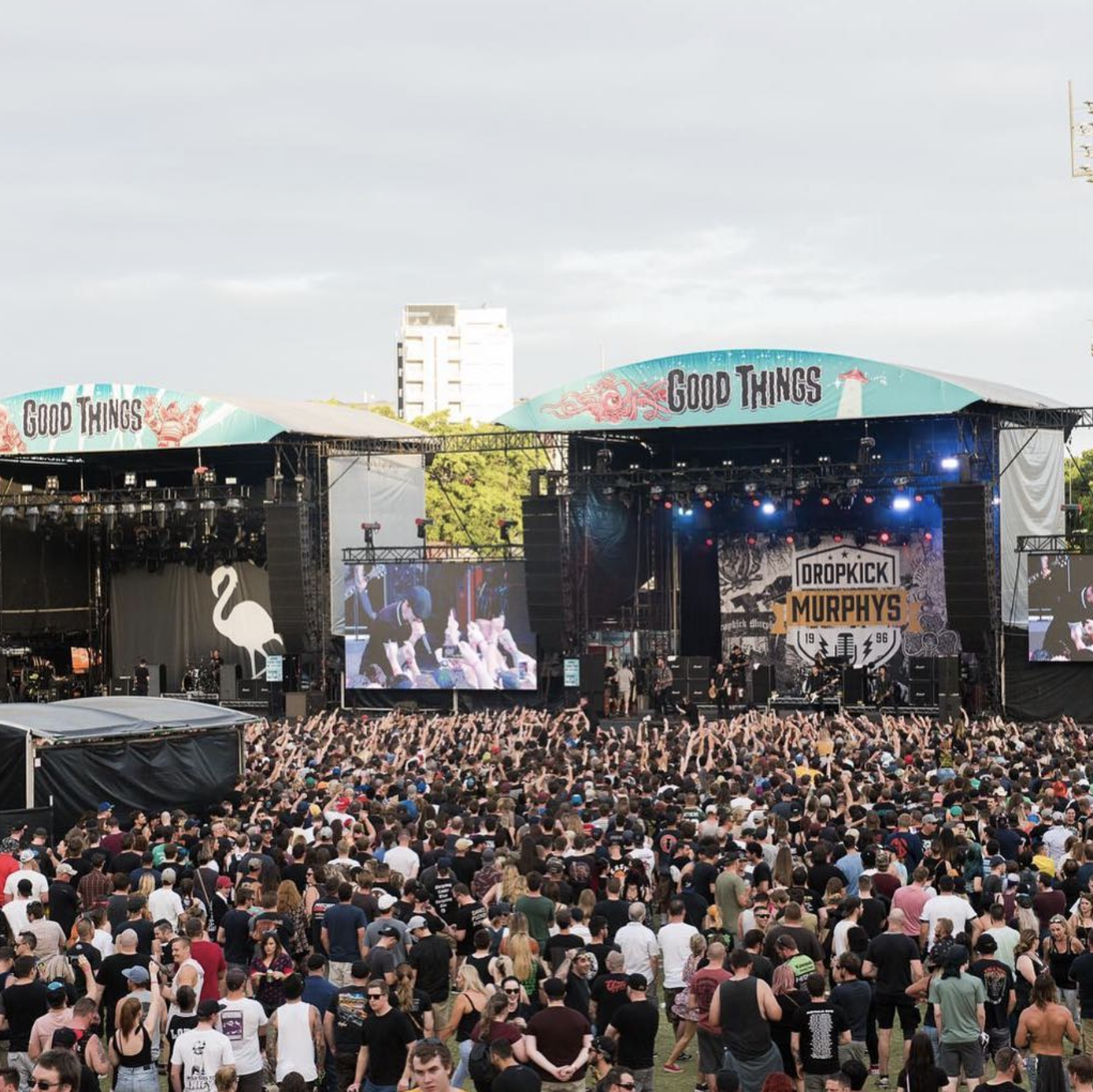 Large outdoor concert with a stage, two big screens showing performers, and a crowd of people in front of the stage, during daytime.