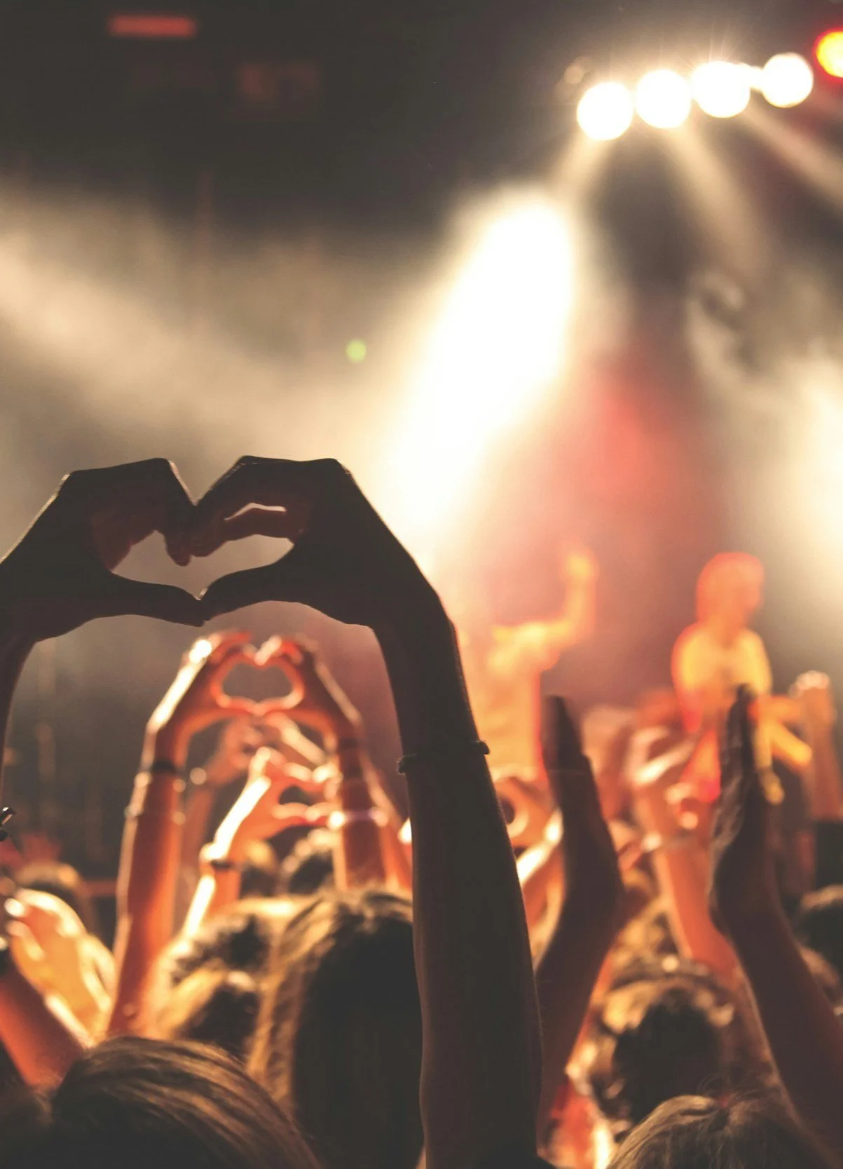 Concert crowd with hands forming heart shapes, stage lights bright and blurred performers in the background.