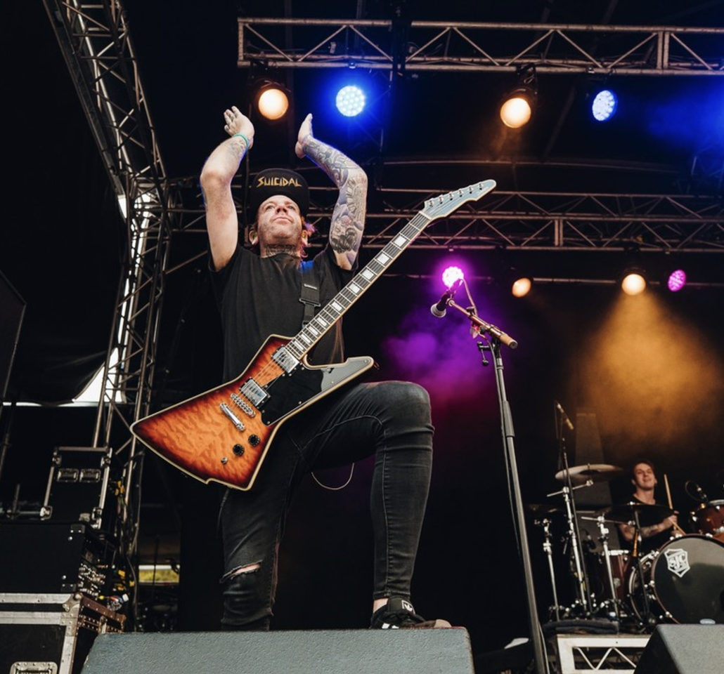 Musician playing electric guitar on stage with lights overhead, wearing a black cap and black clothes, with a drummer in the background.