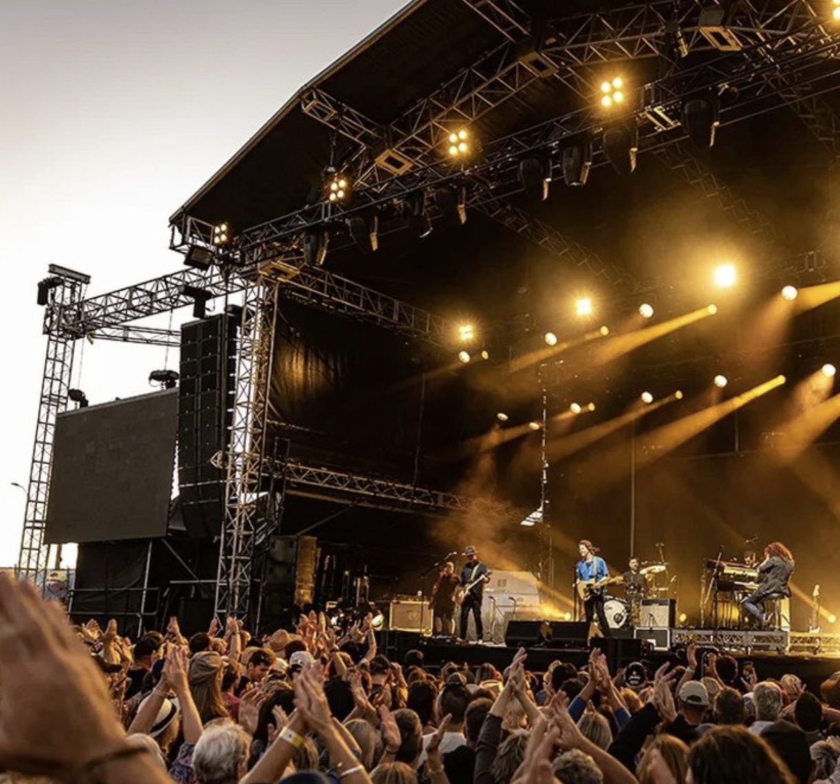 A live outdoor concert with a band performing on stage illuminated by yellow lights, and a large crowd of people facing the stage with hands raised.