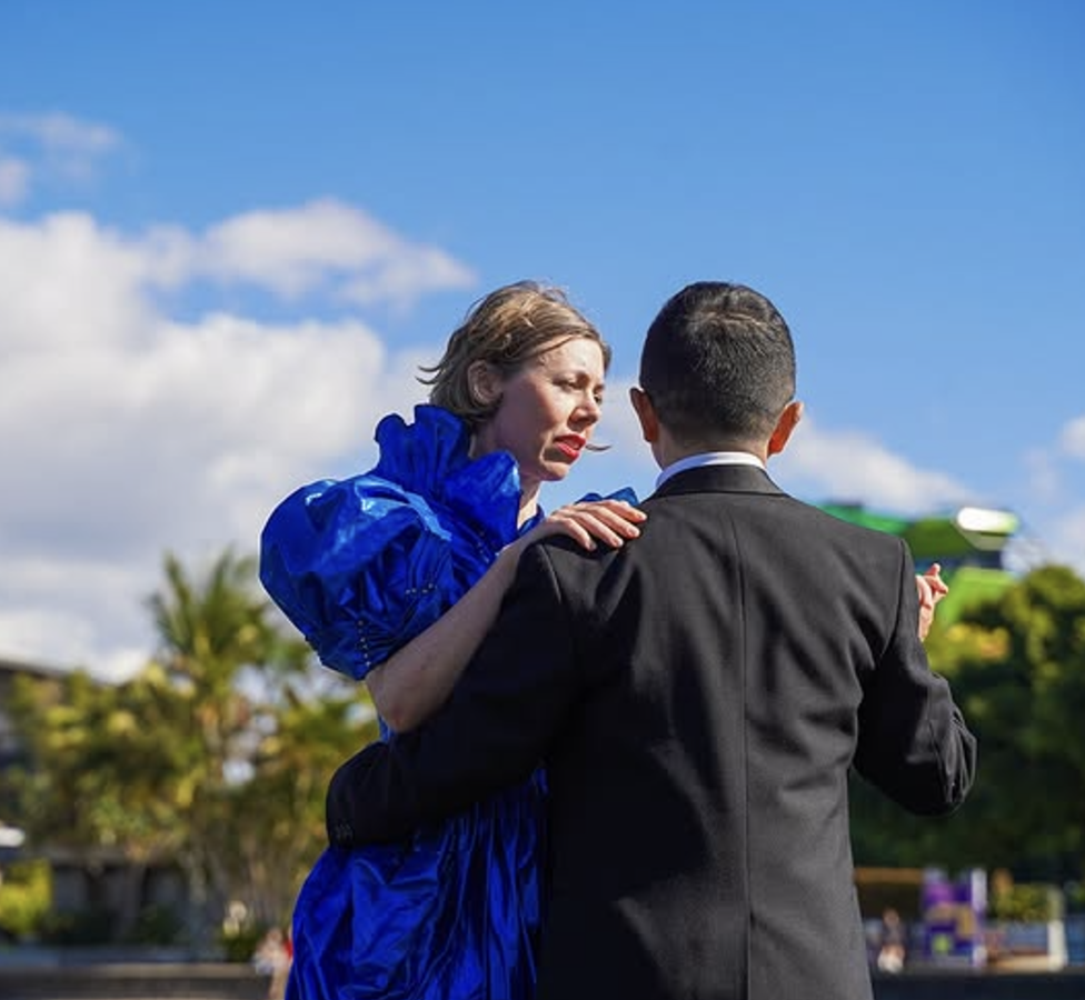 A woman in a bright blue dress with puffed sleeves dancing with a man in a black tuxedo in an outdoor setting with blue sky and clouds.