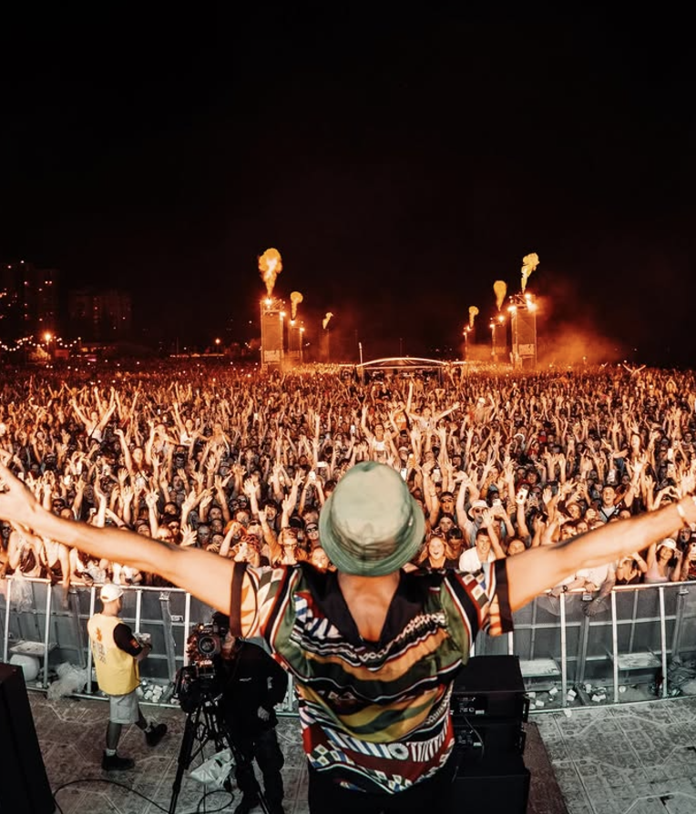 A performer on stage with arms outstretched facing a large crowd at a nighttime outdoor concert with pyrotechnics and bright lights in the background.