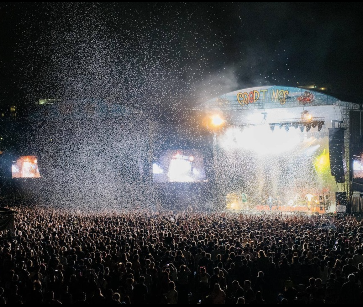 Crowd at a concert or festival at night with a large stage, colorful lights, and a fireworks display in the night sky.
